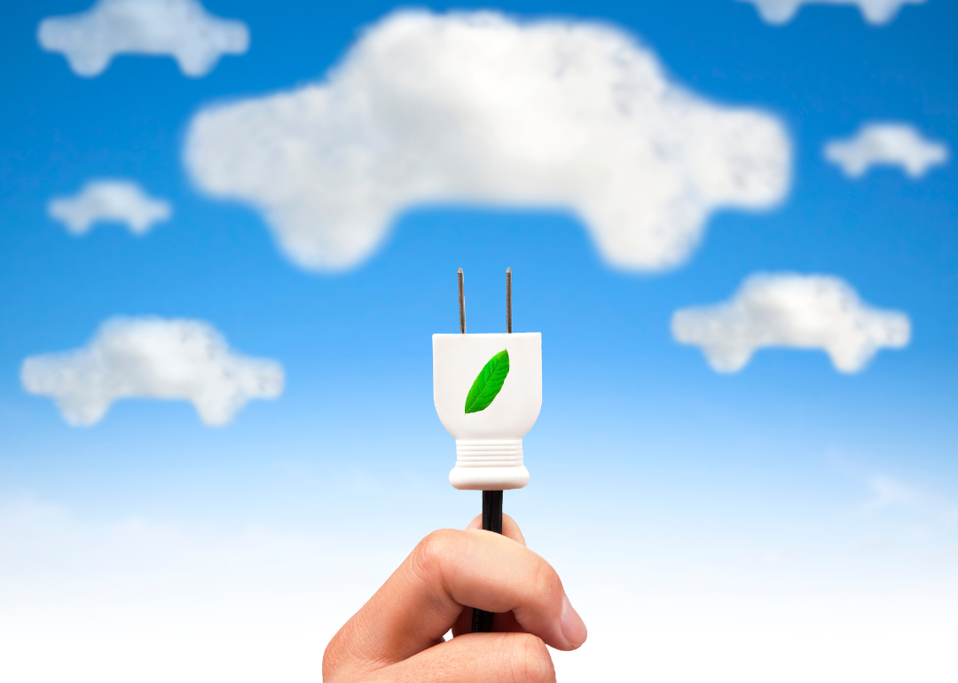 A conceptual image of clouds shaped like cars and a hand holding up an electric plug with a green leaf on it.