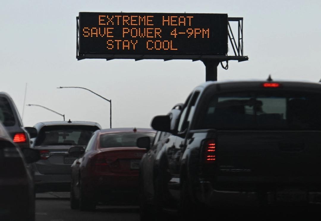 Cars drive under a sign reading “Extreme heat, save power 4-9 pm, stay cool.”