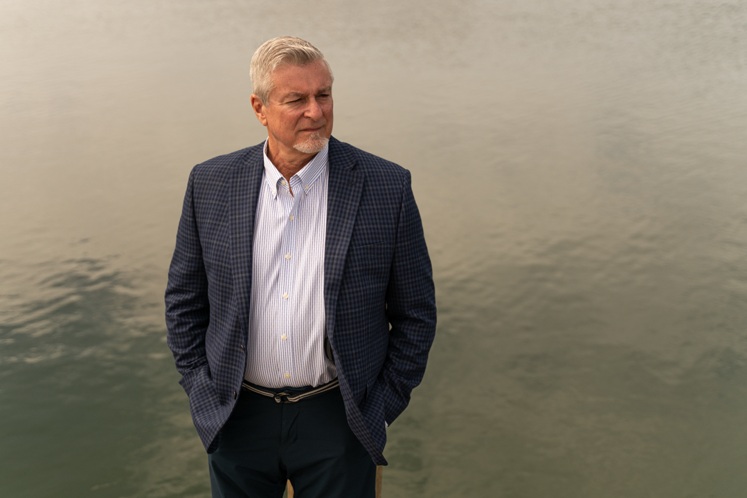A man in a sport coat stands in front of a body of water.