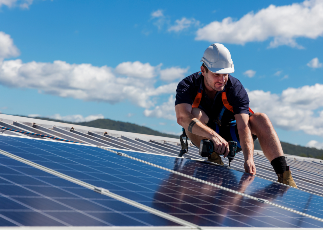 A worker in a white hardhat installing solar panels on the roof of a home.