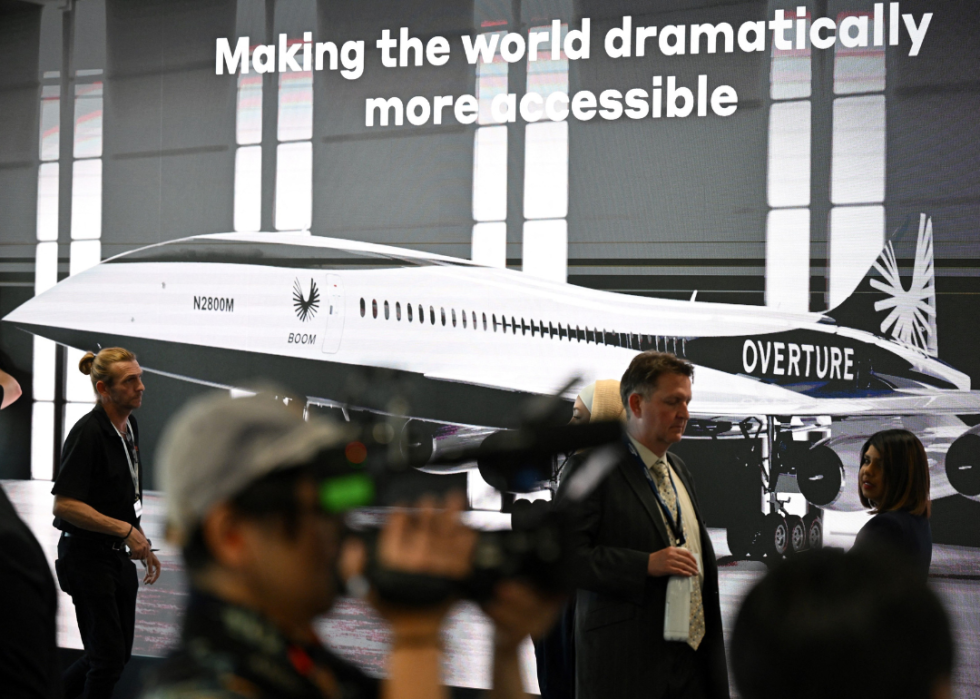 Passengers in an airport walk by a large advertisement for an Overture airplane