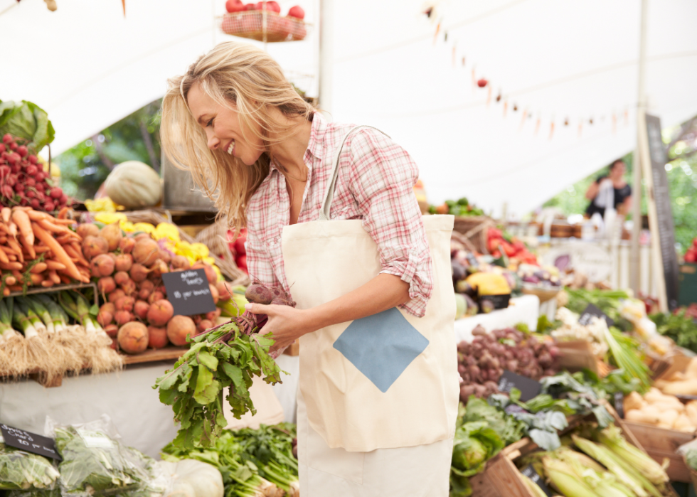 A woman with a bag over her shoulder holds vegetables and smiles at a farmers market