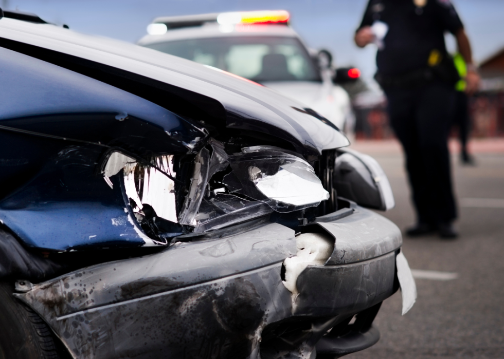 A closeup of a black car that has been in a collision, with a police officer walking towards the vehicle in the background