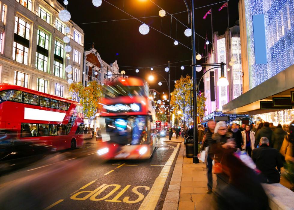 Red double decker buses drive along a busy London street full of people walking on the sidewalk.