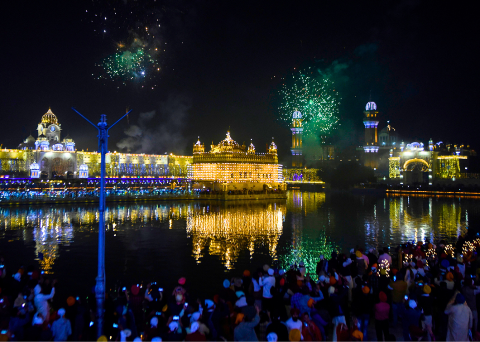 At night, a crowd watches a firework display from across a pond in front of a temple.
