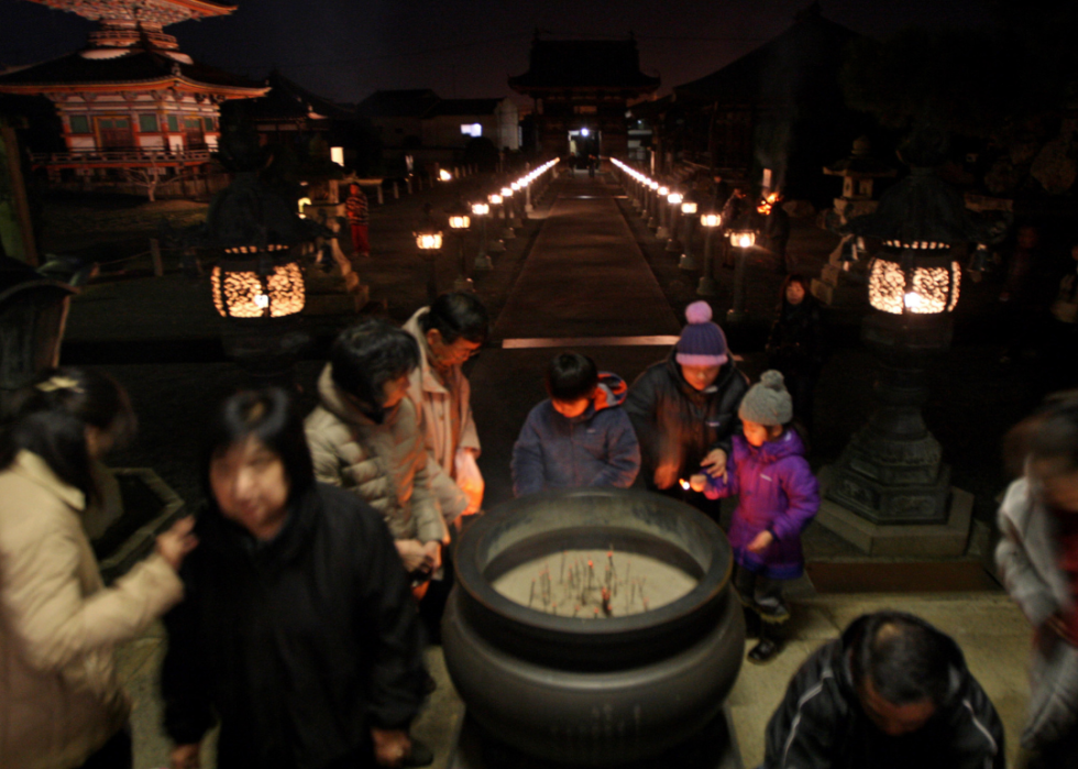 A small group of people with their heads bowed around a vessel filled with sand and lit matches outside temples at night.