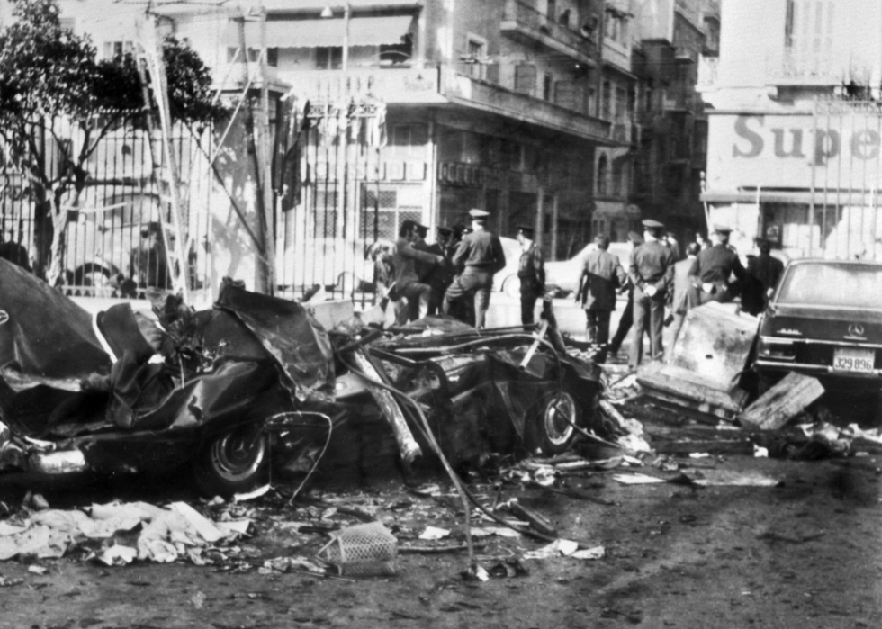 Police stand in the distance with a demolished car in the foreground on a city street.