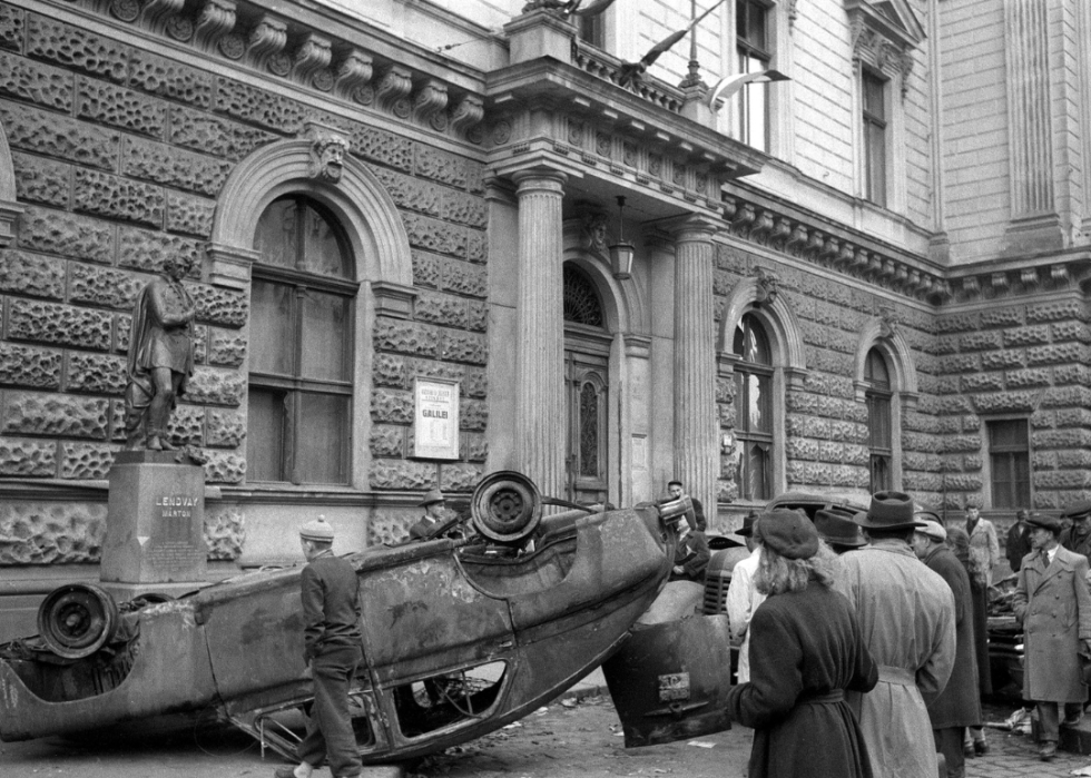 A crowd of people gather around a car that has been turned upside down and destroyed in front of a building with a statue of Lendvay Marton.