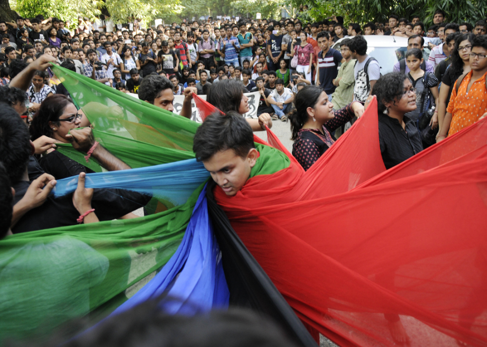 Crowds gather in the street with a group of people wrapping one person in green, blue, red, and black fabric. 