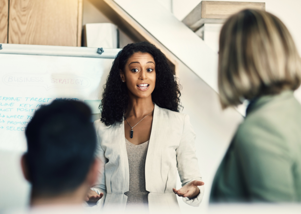 A woman stands in front of a white board facing two people in an office.