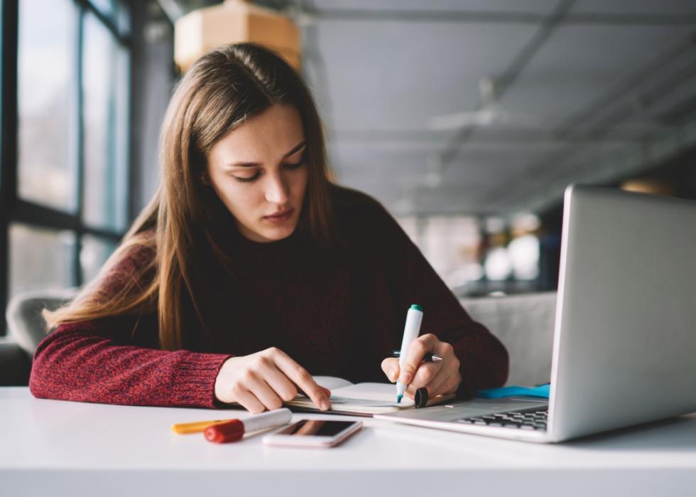 A young woman taking notes by hand while seated in front of a laptop.