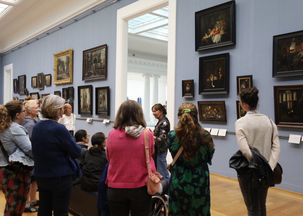 A group on a tour of the museum's collection viewing a room with artwork on pale blue walls. 