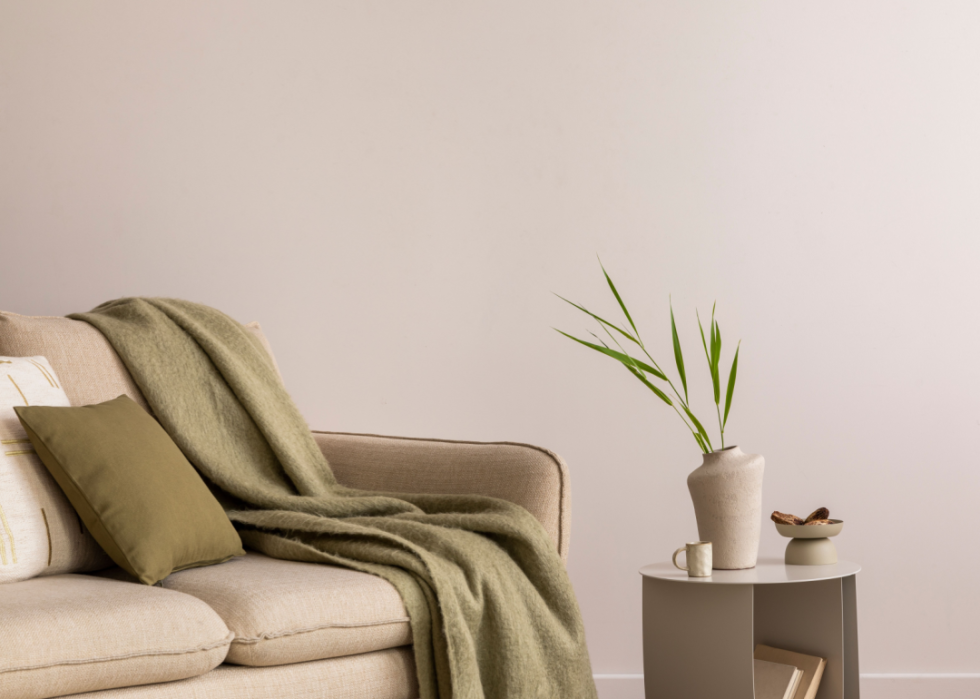 A beige couch with a darker gray blanket is in a minimalist room with a simple table and vase.