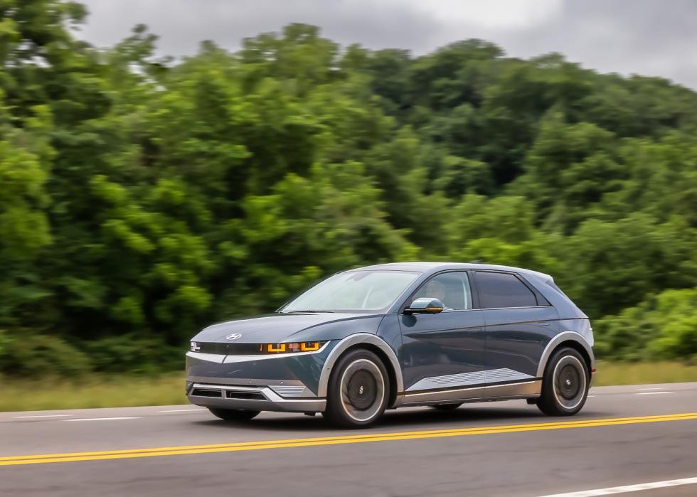 Photo of Dark Hyundai Ioniq 5 driving on road with trees in background. 