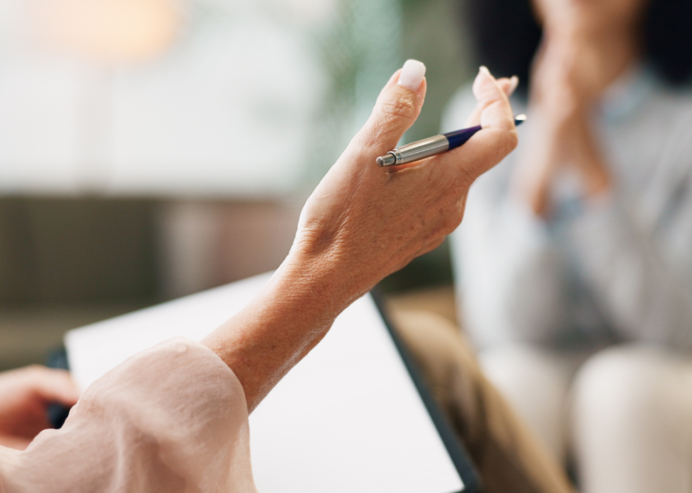 The hand of a therapist in focus, with a woman seated across from her in an office.