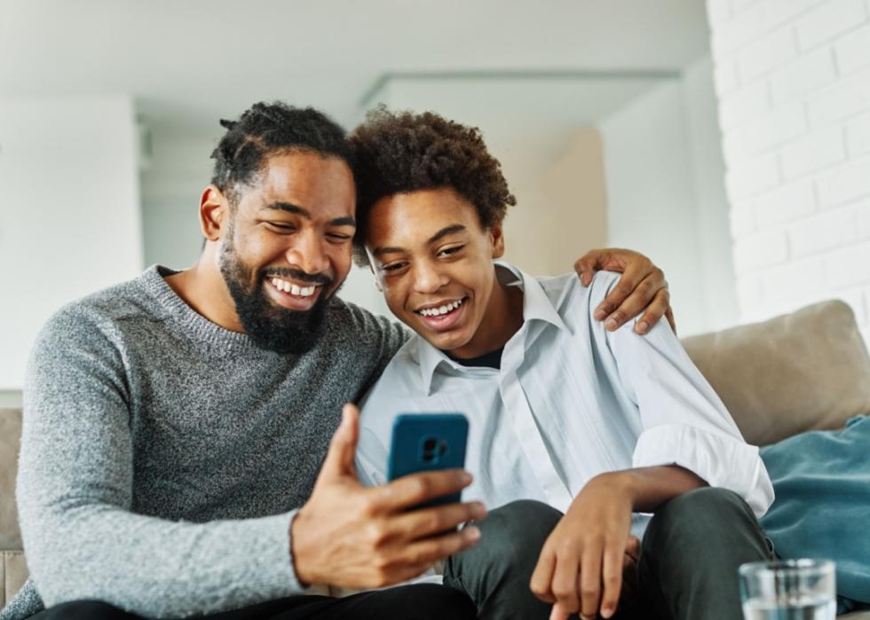 Father and son look at smartphone while sitting on couch.