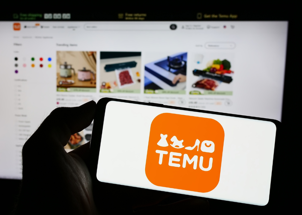 Person holding cellphone with logo of US online shop company Temu on screen in front of business webpage. 