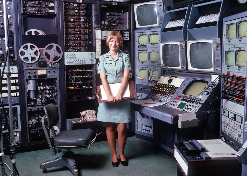A woman in an Army uniform stands in front of a bays of televisions and audio devices