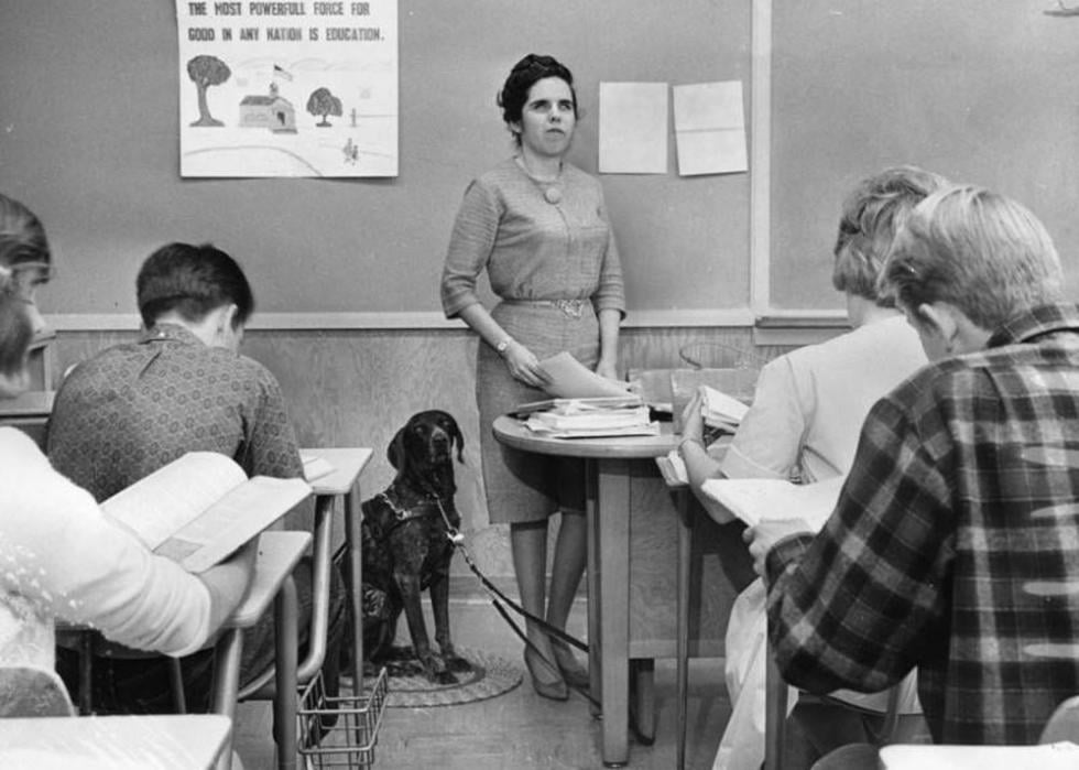 Black and white photo of a blind teacher and her dog in front of a class