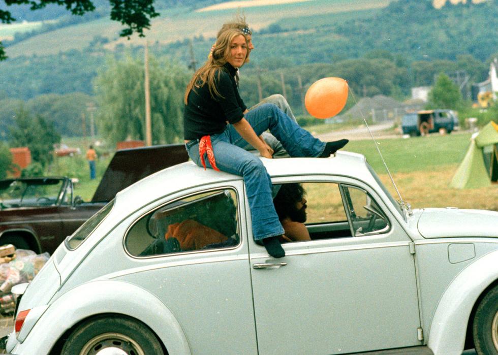 A couple sitting on the roof of a white VW bug driving down the road