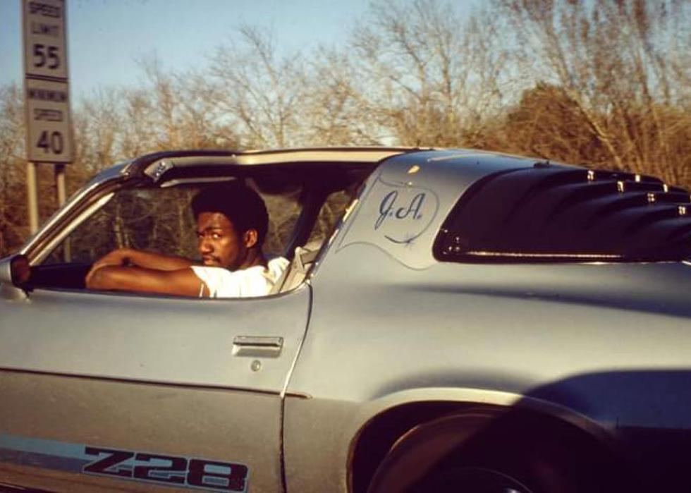 A looking back at the camera while behind the wheel of a silver Z28 Camaro
