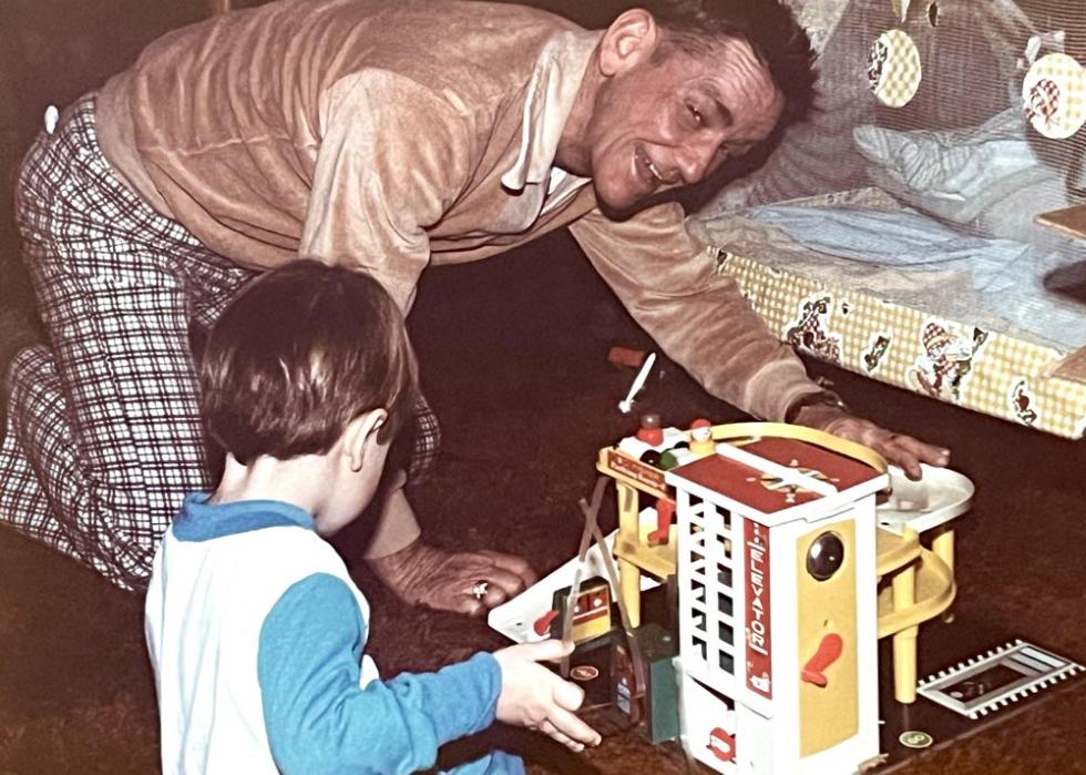An older man on the floor playing with a kid and a fisher price parking garage. 