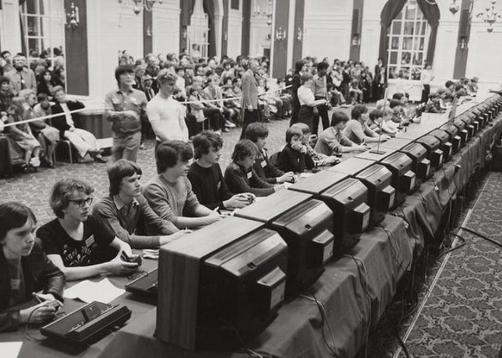 Black and white photo of a row of kids playing space invaders in front of an audience