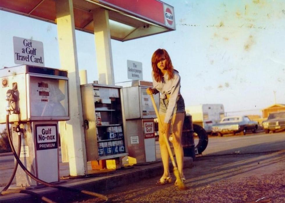 A color photo of a woman sweeping the area in front of the gas pumps at a gas station