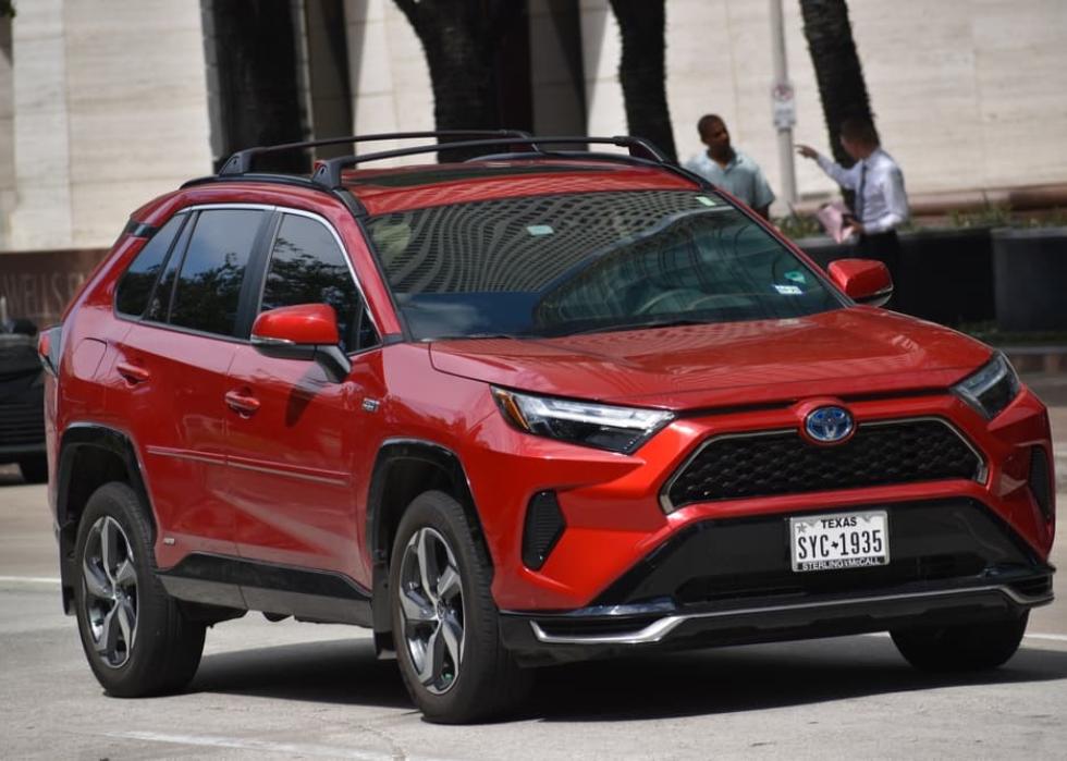 A portrait of a red Toyota RAV4 Prime SUV cruising in a downtown financial district.