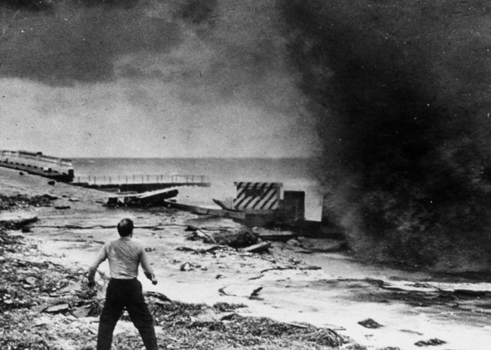 A man stands beneath what looks like a massive plume of dark smoke on a beach surrounded by wreckage.