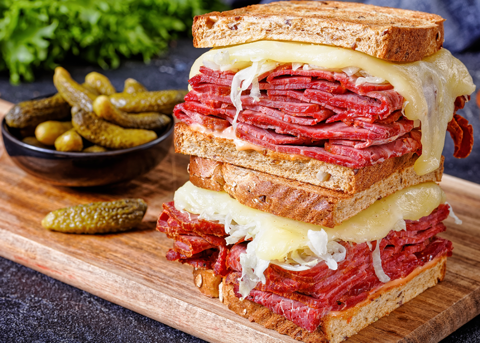 Reuben sandwich with rye bread on a board with pickles in a bowl.