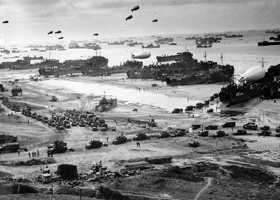A large number of ships in front of Omaha Beach delivering troops, equipment and vehicles lining up to leave the beach, barrage balloons are up in the sky.