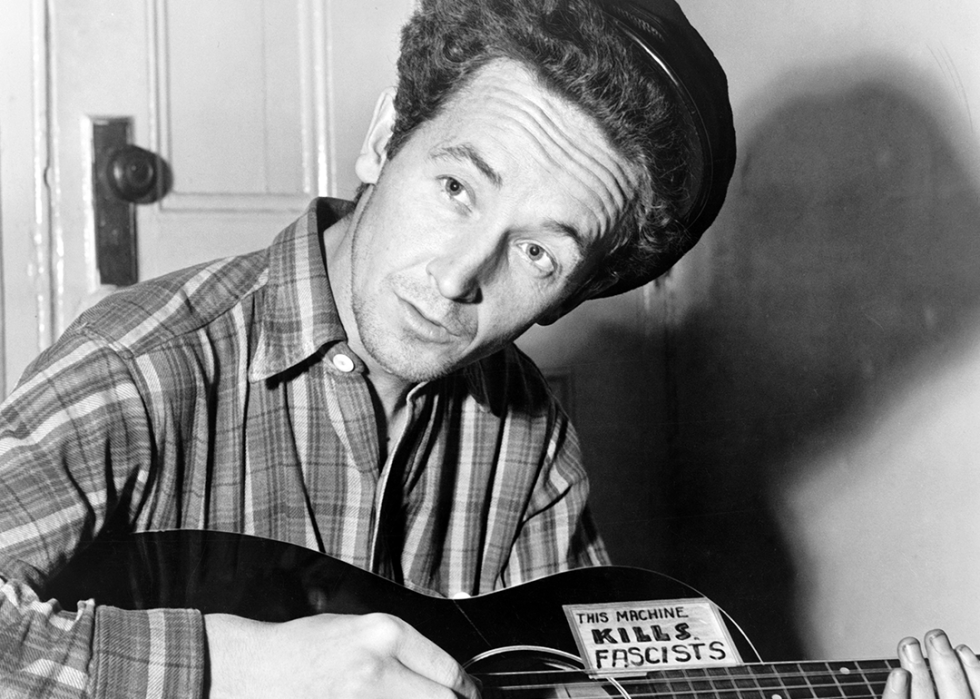 Woody Guthrie poses for a portrait with his guitar which has a sign on it that reads "This Machine Kills Fascists”.
