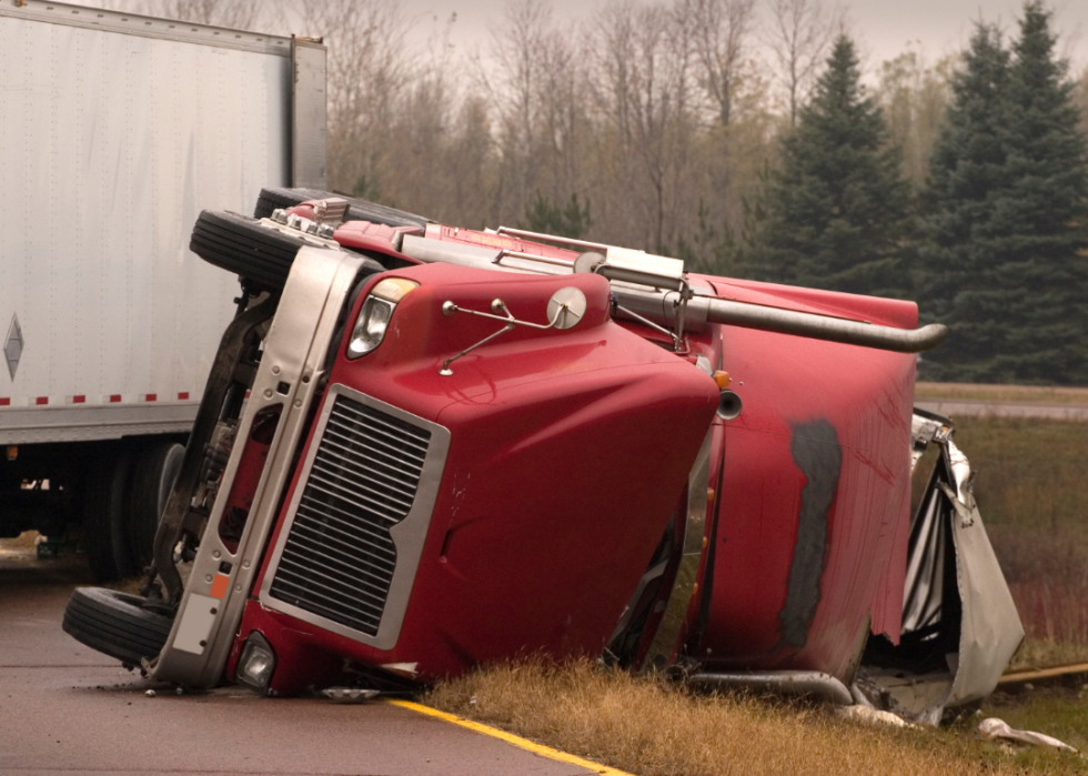 The cab of a red semi truck is tipped over by the side of a road
