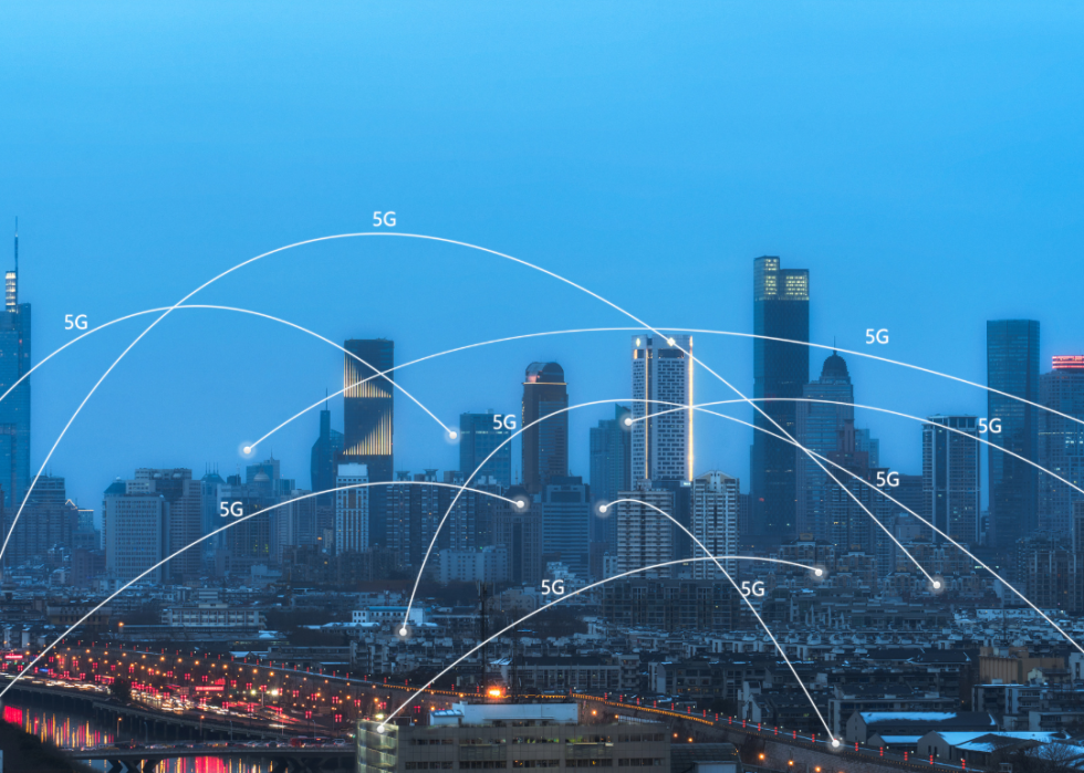 A brightly lit city skyline with skyscrapers and a overlaying digital illustration of a network of blue 5G lines radiating outward and upward from the city.