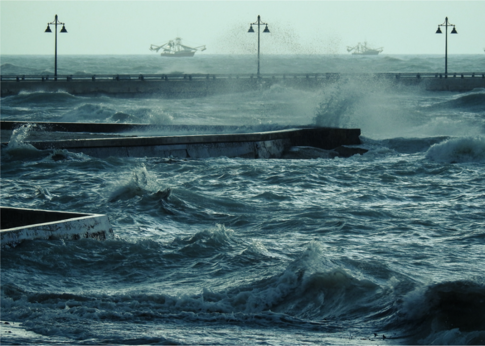 Waves crash against structures and lamp posts with two large boats in the distance.