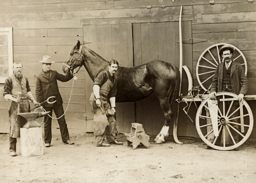 Farrier shoeing a horse with blacksmith and two other men standing nearby.