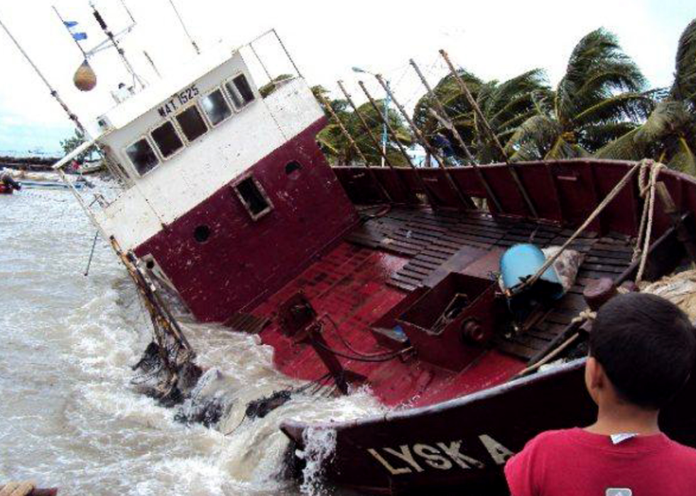 A kid looks out at a fishing boat that is nearly on its side near the shore.