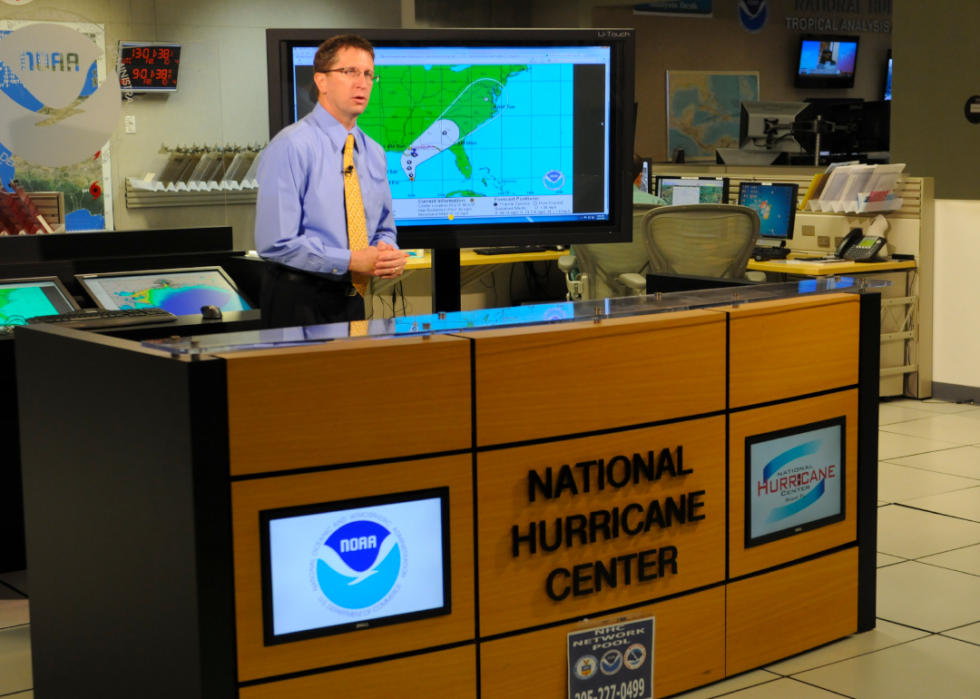 A meteorologist at the National Hurricane Center stands behind a desk and in front of a map on a screen.