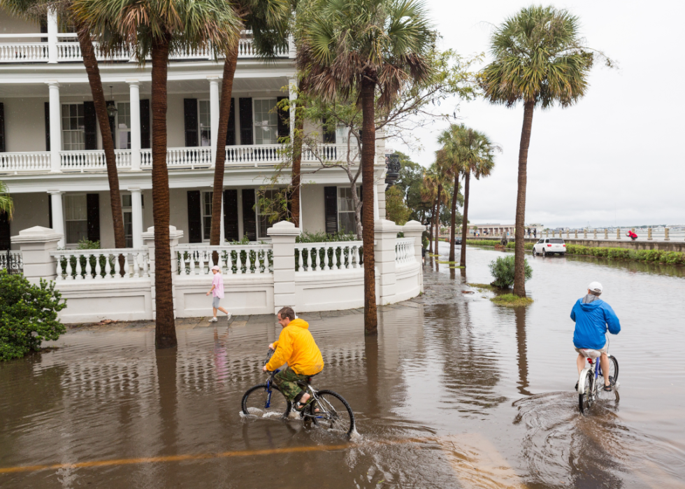 Two people ride their bikes and one person walks down a flooded street lined with palm trees in front of a stately home.