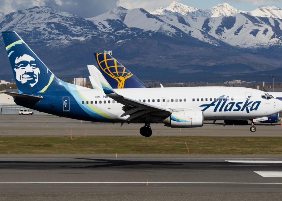 Alaska Airlines aircraft in the foreground, landing on a runway with mountains in the background.  Airline's logo featuring a head of a man on the tail. The mountains in the background are snow-capped. 