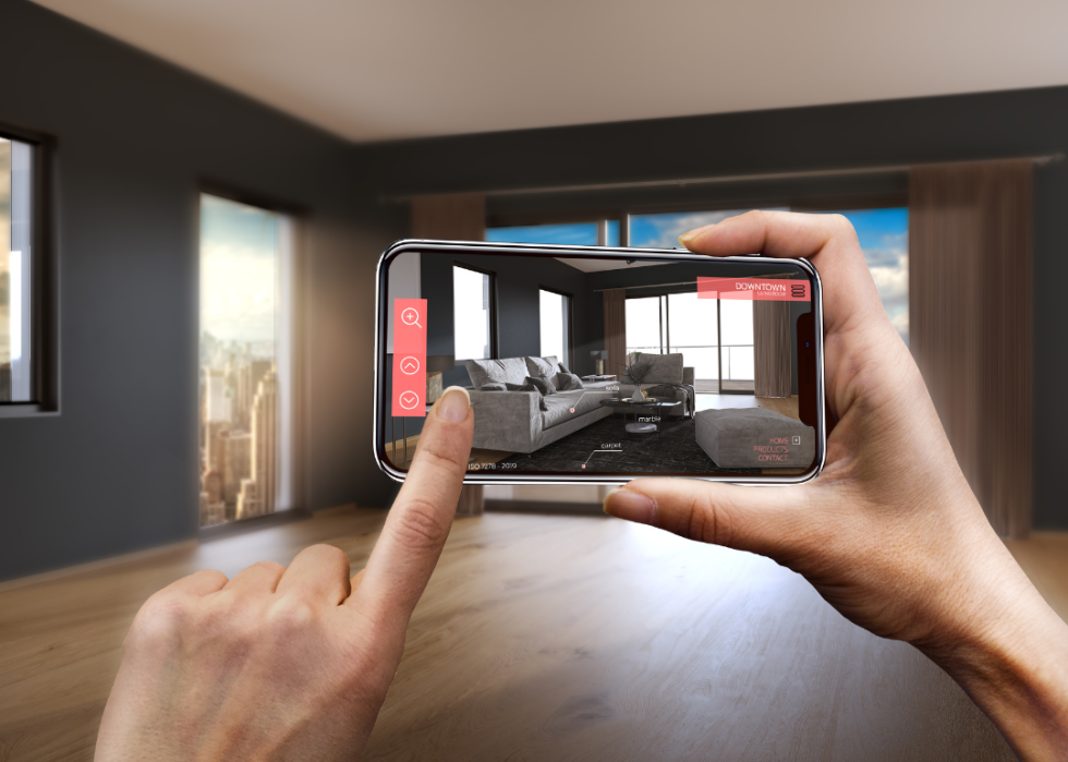 A close up of two hands holding a cell phone toward an empty room with the screen showing what the room would look like with added furniture.