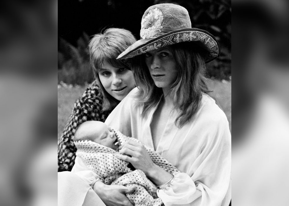 Angela poses with David Bowie, holding their newborn wrapped in a knit blanket.