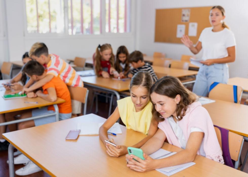 A lively classroom scene with students engaged in group activities. In the foreground, two girls are seated at a desk, closely looking at their smartphones with focused expressions.