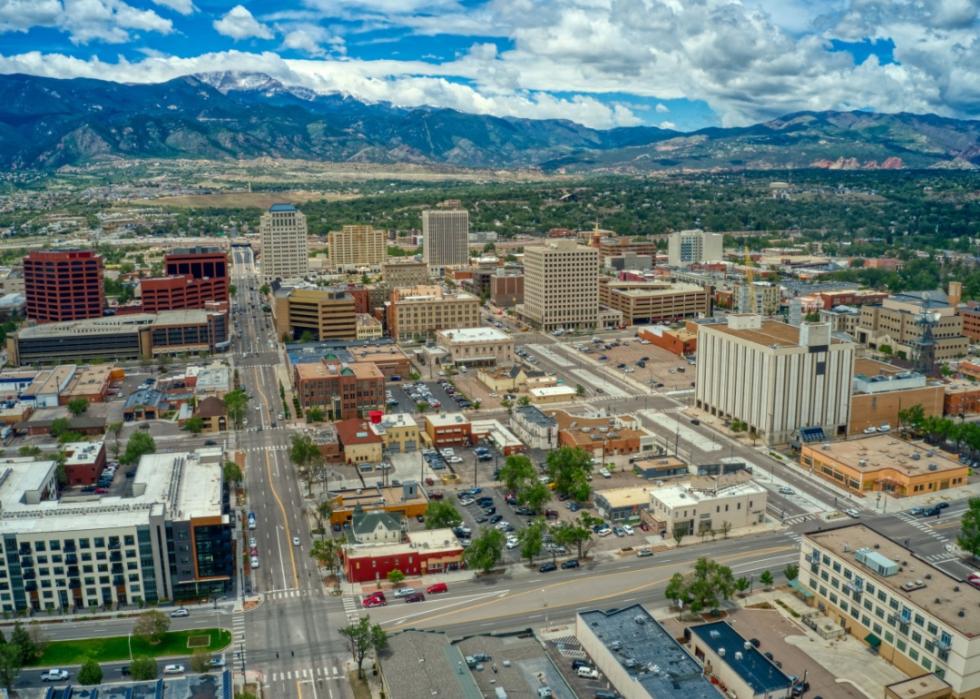 An aerial view of a city with a mountain range in the background. The city has a mix of low and high-rise buildings, with a grid-like street pattern.