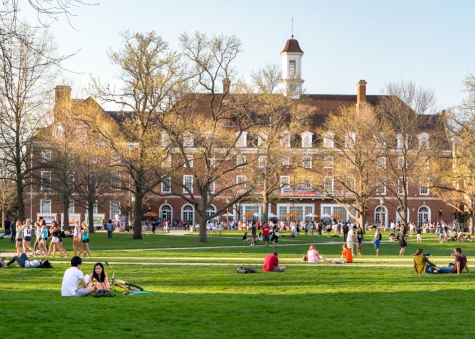 A large green lawn where people are lounging, sitting in groups. Some are sprawled on the grass with books while others are walking or cycling.In the background is a large, classic brick building with white-trimmed windows and a central cupola. 