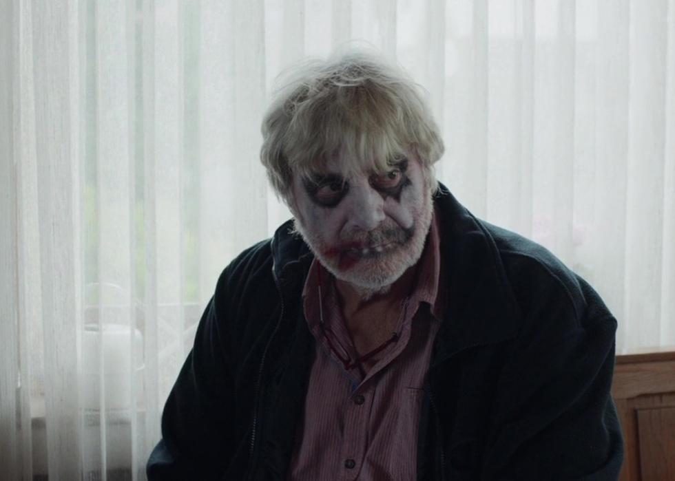 An older man with gray hair has messy clown makeup on his face.