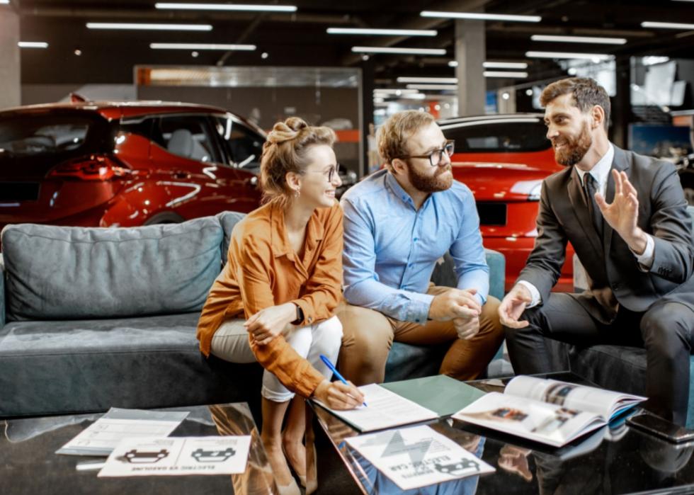 Three individuals seated on a couch in what appears to be a car showroom. A couple, casually dressed, is sitting together, engaged in a conversation with a man in a suit. On the table in front of them are documents, brochures, and a pen.