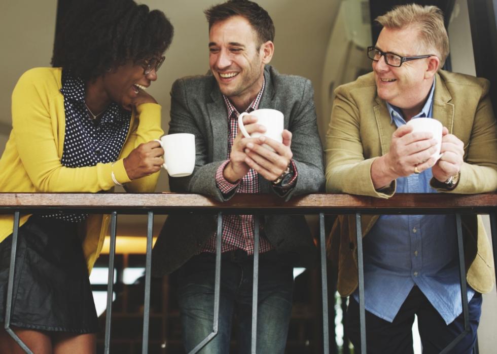 Two male coworkers having coffee with a woman coworker while leaning on a railing.