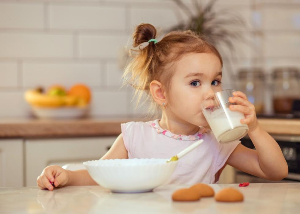 A young girl sitting at a kitchen table, drinking from a glass of milk. In front of her there is a white bowl and a spoon. Her hair tied up in a cute, messy style with a small ponytail. 